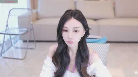 Media: Video of a young East Asian woman with long black hair, fair skin, and delicate features, sitting in a modern living room with beige couches, a glass coffee table, and a minimalist decor.