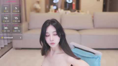 Media: A video of a young, slim Asian woman with long, straight black hair, wearing minimal makeup, sitting topless in a modern living room with a beige couch, a blue blanket, and a blurred background.