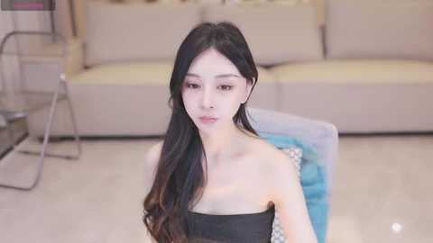 Media: Video of a young Asian woman with long black hair, fair skin, and a slim build, wearing a strapless black dress, sitting on a blue chair in a modern, beige-lit living room with a beige sofa in the background.