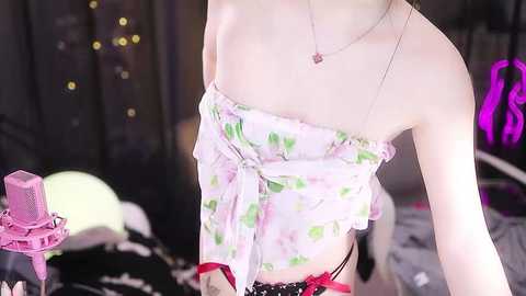 Media: Video of a slender, light-skinned person with pale skin, wearing a floral off-shoulder top, black lace panties, and red suspenders, holding a pink microphone, against a dark backdrop with fairy lights and a purple neon sign.