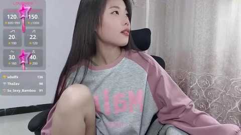 Media: Video of an East Asian woman with long black hair, wearing a grey and pink sweatshirt, sitting on a black chair, looking contemplative. Background includes a weather app and floral curtains.