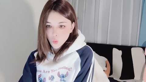 Media: Video of a young woman with light skin, straight brown hair, and large brown eyes. She wears a white and navy hoodie with a cartoon print and looks slightly confused. Background features a bed with white and black pillows.