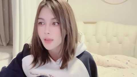 Media: Video of a young Caucasian woman with straight brown hair, wearing a dark hoodie with white sleeves, lying on a bed in a cozy bedroom with light-colored walls and a tufted headboard.