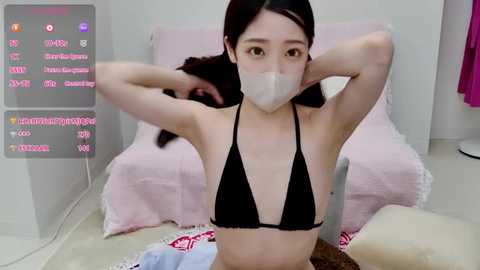 Media: A video of a young East Asian woman with long black hair, wearing a black bikini, face mask, and a pink towel, sitting on a white floor.