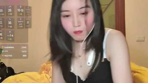 Media: A video of an East Asian woman with long black hair, wearing a black top with white lace, listening to music with white earphones. Background features a yellow bedspread and a digital clock displaying time and weather.