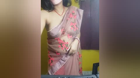 Media: A video of a woman in a see-through pink sari with red floral embroidery, her dark hair visible, standing in a room with yellow and maroon walls.