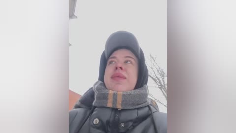 Media: Video of a woman in a dark winter coat and hat, looking up with a pensive expression, standing in a snowy, foggy outdoor environment.