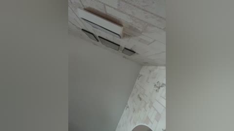 Media: Video of a ceiling with a rectangular air conditioning unit and a marble-tiled wall in a light gray, minimalist room. The image captures a clean, modern interior design with smooth, uncluttered surfaces.