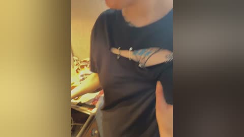 Media: Video of a person wearing a black T-shirt with a cutout design and a colorful pattern on the shoulder, standing next to a kitchen counter with a cluttered surface.