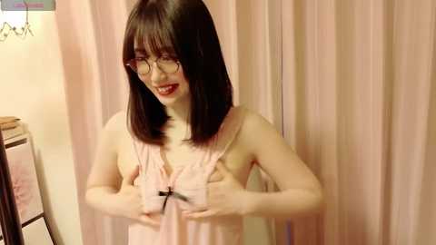 Media: Video of a young Asian woman with straight, shoulder-length black hair, wearing glasses and a pink sleeveless top, playfully holding her breasts in front of light pink curtains.