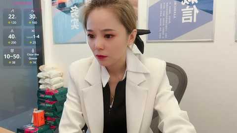 Media: Video of an Asian woman with short, light brown hair, wearing a white blazer over a black top, sitting at a desk in a modern office.