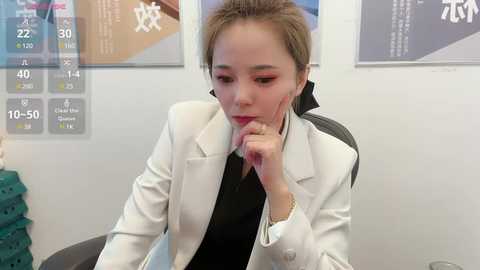 Media: Video of an Asian woman in a white blazer, black shirt, and red lipstick, with short brown hair, hand to chin, looking thoughtful in a modern office setting.