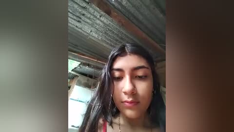 Media: Video of a young Latina woman with long black hair, wearing a red tank top, standing in a rustic, partially constructed wooden building with exposed beams and rough surfaces.