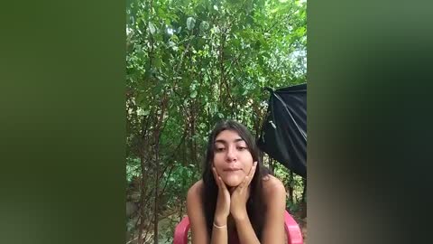 Media: Video of a young woman with long dark hair, leaning on her hands, smiling in a lush green forest.