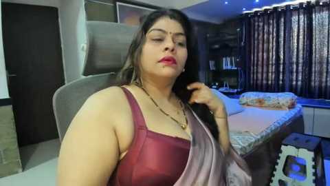 Media: Video of a South Asian woman with long dark hair and medium skin tone, wearing a burgundy bra and pink saree, sitting on a couch in a modern living room with dark curtains.