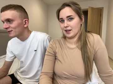 Media: Video of a young Caucasian couple sitting on a bed. The man, with short brown hair, wears a white t-shirt. The woman, with long blonde hair, wears a beige top. Background shows a wooden door and beige walls.