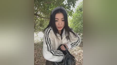 Media: Video of an Asian woman with long black hair, wearing a white Adidas jacket with black stripes, standing outdoors in a green, leafy area.