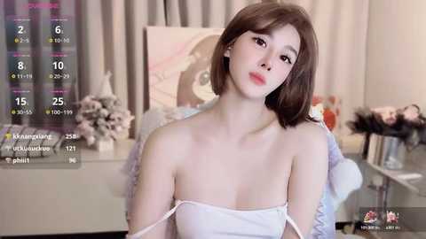 Media: Video of a young, fair-skinned woman with shoulder-length brown hair, wearing a white, strapless top, seated in a softly lit bedroom. Background includes a flower arrangement, a TV, and a framed picture.