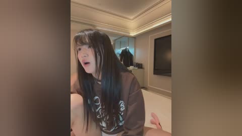 Media: A video captures an Asian woman with long black hair, wearing a dark T-shirt, kneeling in a modern, dimly lit living room with beige walls and a flat-screen TV.