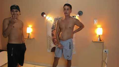 Media: Video of two shirtless teenage boys in a dimly lit room. One wears a backwards baseball cap, black shorts, and holds his hands to his mouth; the other wears light blue shorts, smiling, with a reflective surface behind them.