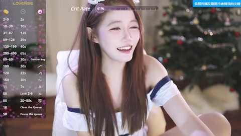 Media: A video of a smiling East Asian woman with long brown hair, wearing a white off-shoulder top and a festive headband, sitting in front of a decorated Christmas tree.