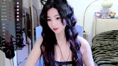 Media: Video of an Asian woman with long black wavy hair, wearing a black lace top, sitting in a bedroom with plush toys, a mirror, and a recording overlay.