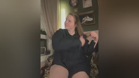 Media: A candid video of a plus-sized woman with light skin and long brown hair, wearing a black bodysuit, adjusting her hair in a cozy living room with green walls, framed art, and a floral-patterned couch.