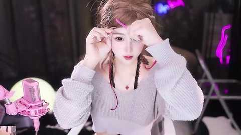 Media: Video of a pale-skinned woman with brown hair, adjusting pink hair clips on a light gray knit sweater, against a dark stage with neon lights.