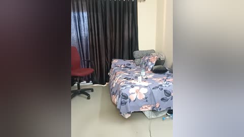 Media: Video of a simple, cluttered bedroom with a bed covered in a floral-patterned duvet, gray pillows, a red chair, and a dark curtain backdrop.