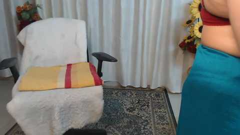 Media: Video of a woman in a teal dress standing beside a white armchair draped with a yellow blanket, in a room with white curtains and floral arrangements.