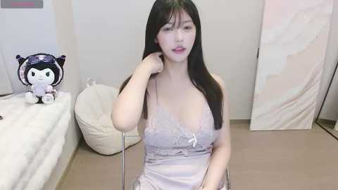 Media: Video of an East Asian woman with long black hair, wearing a revealing light purple lace lingerie top, sitting on a beige bean bag chair in a minimalist room with a plush toy on a white bed.