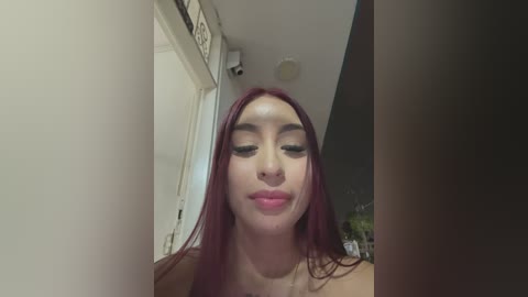 Media: Video of a young woman with long, straight, dark red hair, wearing minimal makeup, and a nose ring. She has a light skin tone and is indoors, looking slightly to the side.