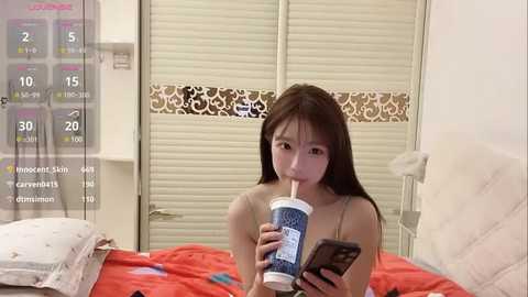 Media: Video of an Asian woman with long brown hair, holding a blue and white straw cup, standing in a bedroom with closed blinds and a bed with red sheets.