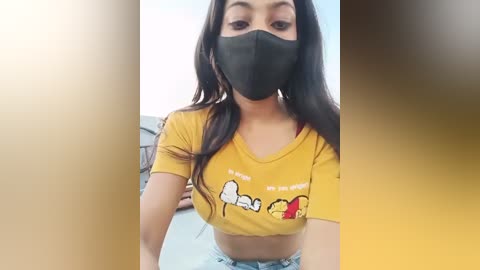 Media: A video of a young woman with long dark hair, wearing a yellow crop top with cartoon characters and a black face mask, sitting indoors.