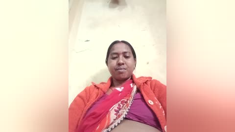 Media: Video of a South Asian woman with dark skin, wearing an orange jacket over a red sari with a white pattern, lying in a narrow, light-colored bed.