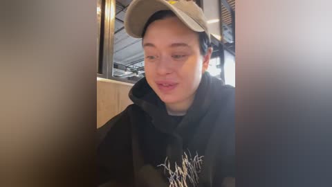 Media: Video of a young man with short dark hair, wearing a beige cap and a dark hoodie, sitting in a dimly lit indoor setting.