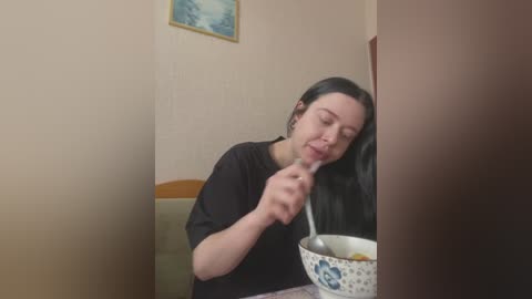 Media: Video of a fair-skinned woman with long black hair, wearing a black shirt, eating cereal from a blue-patterned bowl at a table in a beige room with a framed painting on the wall.