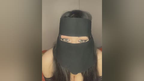 Media: A video of a woman with long, straight black hair, wearing a black mask with eye slits, standing against a plain beige background. She has light skin and is wearing a red top.