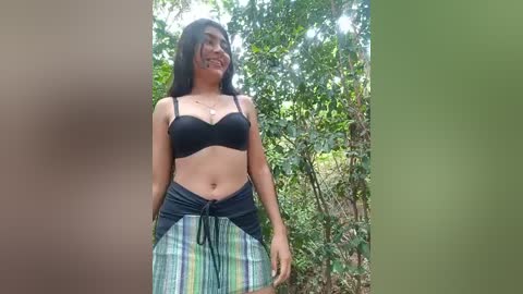 Media: Video of a young woman with long black hair, medium skin tone, wearing a black bra and plaid skirt, standing in a lush, green forest.