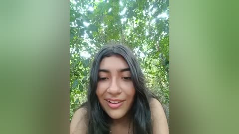 Media: A video of a young woman with long, dark hair and medium skin tone, smiling. She is outdoors, surrounded by lush green foliage, with sunlight filtering through the trees.