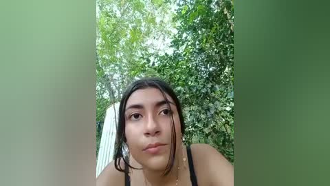 Media: Video of a young woman with light brown skin, straight dark hair, and brown eyes, wearing a black tank top, posing outdoors with lush green trees in the background.