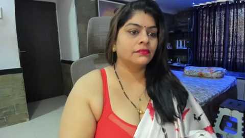 Media: Video of a South Asian woman with medium skin tone, long black hair, and a red top, sitting on a beige chair in a dimly lit bedroom with a bed, curtains, and wooden furniture.