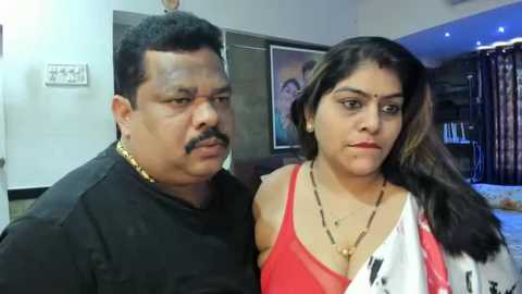 Media: Video of an Indian couple: a middle-aged man in a black shirt and gold chain, and a young woman in a red top, standing indoors.