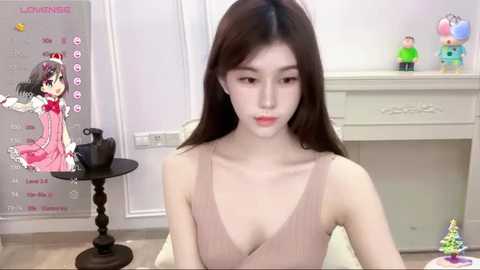 Media: Video of a young East Asian woman with long brown hair, wearing a sleeveless pink dress, sitting in a room with white walls, a black stool, and a white cabinet.