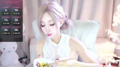 Media: Video of an Asian woman with light skin, blonde hair, and a white lace top, eating noodles in a cozy room with soft, pastel-colored lighting and a plush chair.