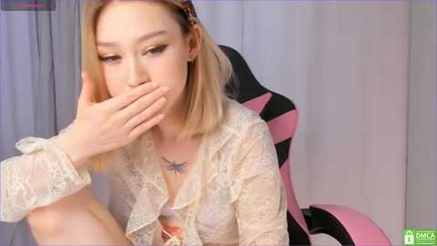 Media: A video of a young Asian woman with light skin and straight blonde hair, wearing a sheer lace blouse, covering her mouth with her hand, seated in a pink and black gaming chair.