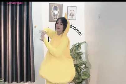 Media: Video of an Asian woman in a yellow Pikachu costume, smiling, in a home setting with potted plants and framed pictures on a white wall.