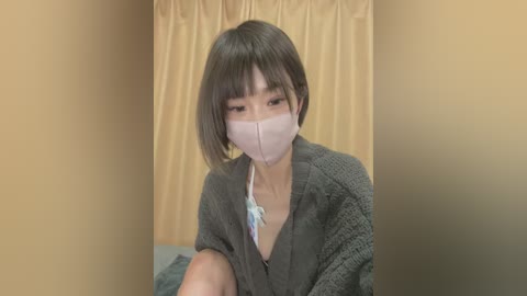 Media: Video of an East Asian woman with short, straight, light brown hair, wearing a gray cardigan, pink mask, and a light blue shirt, seated against a beige curtain backdrop.