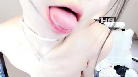 Media: Video of a fair-skinned woman with long black hair, wearing a choker necklace and a white top, licking her lips seductively, taken from a low angle. Background includes a white chair and indistinct objects.