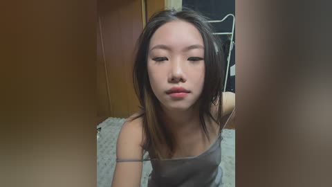 Media: Video of an East Asian woman with straight black hair, wearing a gray spaghetti-strap top, standing in a dimly lit room with beige walls and a patterned carpet.